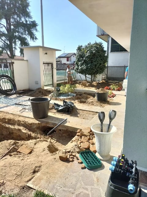 Residential backyard under construction with excavated soil, tools, pots, and young tree plantings visible