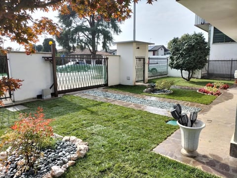 Modern house courtyard with manicured lawn, white metal gates, decorative rocks, potted plants, and autumn trees in background